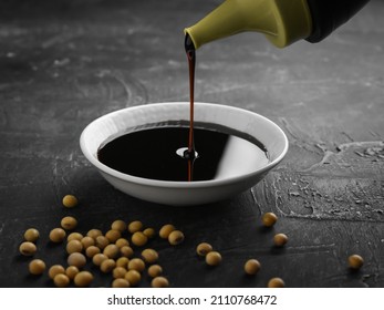 Pouring sweet soy sauce into white ceramic bowl from bottle on black background. soybean seeds in frame
					
