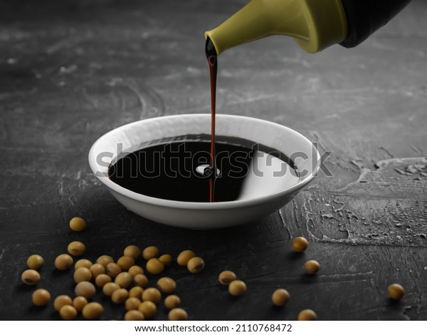 Pouring sweet soy\
sauce into white ceramic bowl from bottle on black background.\
soybean seeds in\
frame\
