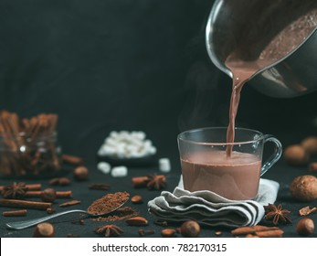 Pouring tasty hot chocolate cocoa drink into glass mug with ingredients on black table. Copy space Dark background. Low key. - Shutterstock ID 782170315