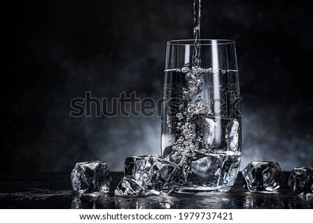 Pouring water from bottle into glass with ice cubes on black background