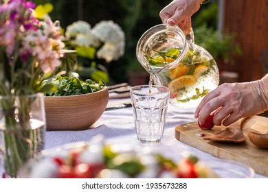 Pouring water with mint herbs and lemon from pitcher into drinking glass. Woman preparing healthy food and drinks