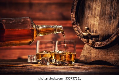 Pouring Whiskey From Bottle To Two Glasses With Ice Cubes, Served On Wooden Planks. Vintage Countertop With Keg And Glasses Of Hard Liquor