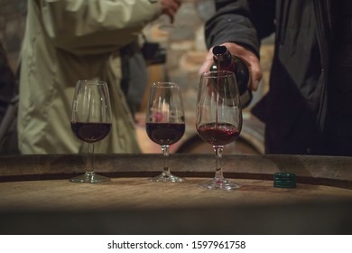 Pouring Wine From A Bottle In Fancy Glasses In A Wine Cellar. People Mingling Around