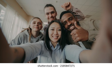 POV Of Asian Girl Holding Smartphone Taking Selfie Photos With Cheerful Multi-ethnic Classmates And Have Fun At University Library Indoors. Woman Pointing Touchsreen And Watching Pictures