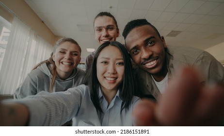 POV Of Asian Girl Holding Smartphone Taking Selfie Photos With Cheerful Multi-ethnic Classmates And Have Fun At University Library Indoors. Woman Pointing Touchsreen And Watching Pictures