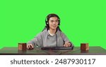 POV of asian guy playing at pc desktop, sitting against full body greenscreen backdrop. Young gamer having fun with players online in gaming competition, wearing headphones at workstation.