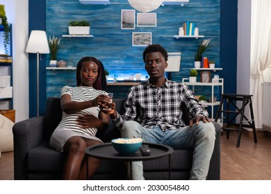 POV Of Black Couple Expecting Child Watching TV In Living Room. African American Young Family Looking At Camera And Television While Pregnant Woman Holding Glass Of Water And Hand On Belly