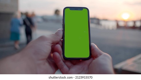 Pov Bokeh Shot Of Man Hands Holding Modern Black Mobile Phone With Blank Green Screen Outdoors. Male Using Smartphone In City, Close Up.