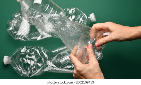 POV Female Hands Crumple And Crush Transparent Plastic Bottle For Recycling. Crumpled Pop Bottles On Green Background. First Point View Preparation Of Recycled Household Trash As Zero Waste