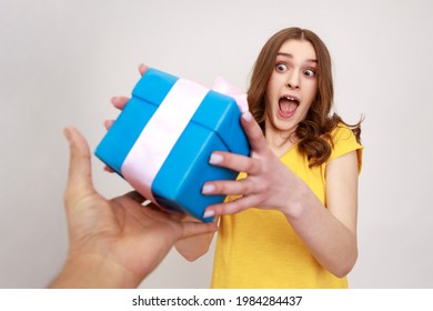 POV Hand Giving Wrapped Blue Gift To Surprised Teenager Girl Looking At Present With Open Mouth, Excitement And Amazement, Congratulation On Birthday. Indoor Studio Shot Isolated On Gray Background.