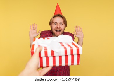 POV Hand Giving Wrapped Gift Box To Guy With Party Cone On Head, Excited Man Raising Hands Shouting For Joy, Looking At Birthday Present Surprise With Delight. Indoor Studio Shot Isolated On Yellow