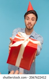 POV Hand Giving Wrapped Red Gift Box To Bearded Man With Party Cone On Head, Guy Wearing Denim Shirt Looking At Birthday Present Surprise With Delight. Indoor Studio Shot Isolated On Blue Background.