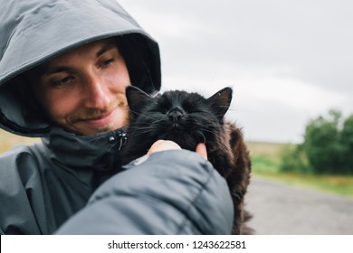 POV Of Hand Of Person Petting And Scratching Cute Adorable Black Kitten Or Cat On Arms Of Young Man In Hood And Jacket. Kindness Towards Animals, Abandoned Pets Rescue