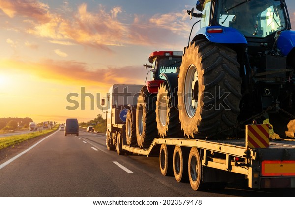 POV heavy industrial truck semi trailer flatbed\
platform transport two big modern farming tractor machine on common\
highway road at sunset sunrise sky. Agricultural equipment\
transportation service