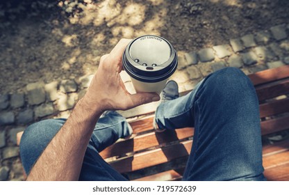 POV Image Of Man Sits On A Bench And Holding Paper Cup Of Coffee At A Summer Day In The Park