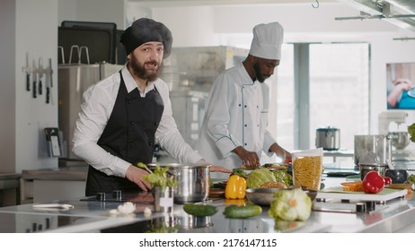 POV Of Male Cook Recording Cooking Show Video On Camera, Explaining Culinary Food Recipe. Young Chef With Uniform Filming Content For Gastronomy TV Program In Professional Kitchen. Tripod Shot.