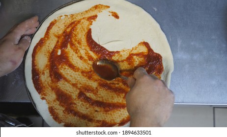 POV male hand of chef spreading tomato sauce on pizza dough using a spoon at kitchen table. Arm of cook applying ketchup on a pastry in metal form at wooden surface. Top view Slow motion