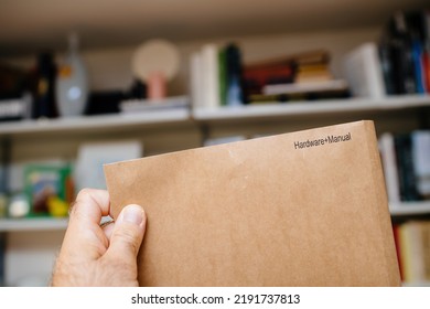 POV Male Hand Holding Paper Envelope With Inscription Hardware Plus Manual - Defocused Room Background With Bookshelves