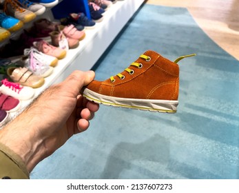 POV Male Hand Holding Small Orange Leather Children Shoe With Multiple Colorful Shoes In Background