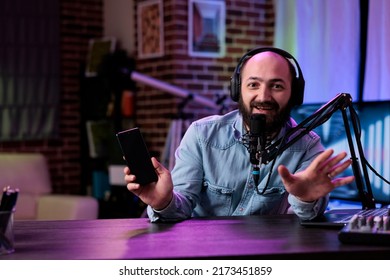 POV of male lifestyle vlogger recommending mobile telephone on podcast, broadcasting live video on social media channel. Cheerful man doing product review with modern smartphone on camera.