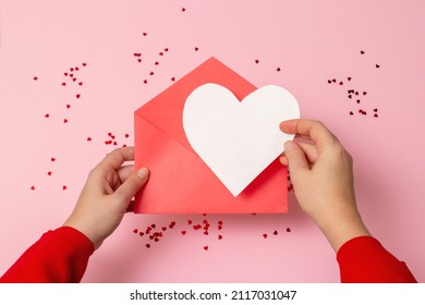 POV Photo From Above: Female Hands In A Red Sweater Holding An Open Pastel Yellow Envelope With A White Paper Heart Over Scattered Sequins On A Light Pink Background With Copyspace