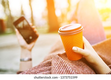 POV Pretty Woman Lying On The Bench In Spring Park And Holding Mobile Phone And Orange Cardboard Cup Of Tea Or Coffee