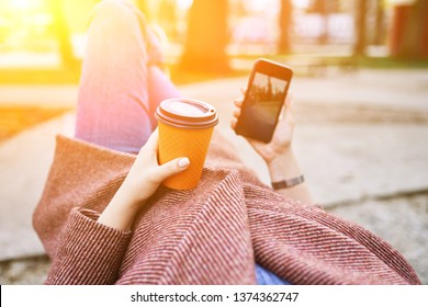 POV Pretty Woman Lying On The Bench In Spring Park And Holding Mobile Phone And Orange Cardboard Cup Of Tea Or Coffee