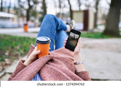 POV Pretty Woman Lying On The Bench In Spring Park And Holding Mobile Phone And Orange Cardboard Cup Of Tea Or Coffee