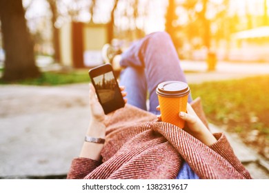 POV Pretty Woman Lying On The Bench In Spring Park And Holding Mobile Phone And Orange Cardboard Cup Of Tea Or Coffee