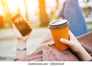 POV Pretty Woman Lying On The Bench In Spring Park And Holding Mobile Phone And Orange Cardboard Cup Of Tea Or Coffee