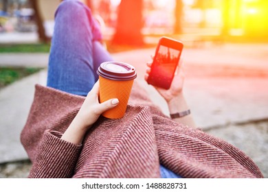 POV Pretty Woman Lying On The Bench In Spring Park And Holding Mobile Phone And Orange Cardboard Cup Of Tea Or Coffee