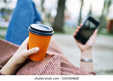 POV Pretty Woman Lying On The Bench In Spring Park And Holding Mobile Phone And Orange Cardboard Cup Of Tea Or Coffee