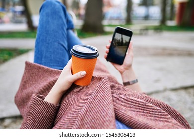 POV Pretty Woman Lying On The Bench In Spring Park And Holding Mobile Phone And Orange Cardboard Cup Of Tea Or Coffee