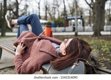 POV Pretty Woman Lying On The Bench In Spring Park And Holding Mobile Phone And Orange Cardboard Cup Of Tea Or Coffee