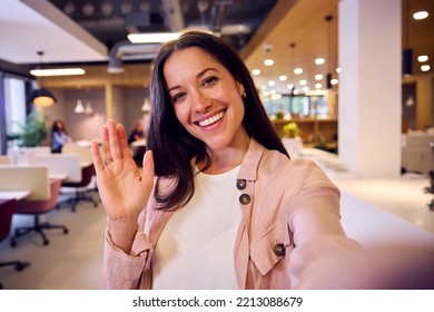 POV Selfie Portrait Of Waving Businesswoman Standing In Modern Open Plan Office