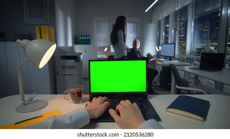 Pov shot of businessman typing on laptop with green screen late in office. Entrepreneurs working at night in office downtown with beautiful night city center view outside the window