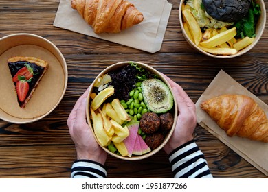 Pov Shot Of Man Holding Hawaiian Vegan Poke Bowl Over The Wooden Table, Takeout Food. French Croissant, Salmon Burger With Black Bun, Blueberry Cheesecake. Close Up, Top View, Copy Space, Background
