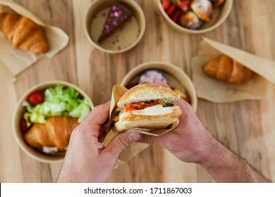 Pov Shot Of Man Holding Italian Panini Sandwich Over The Wooden Table With Takeout Food. French Croissant With Salmon And Chocolate Cheesecake. Close Up, Top View, Pov, Copy Space, Background.