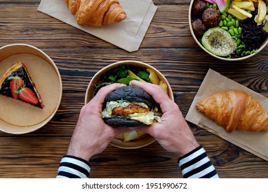 Pov Shot Of Man Holding Salmon Burger With Black Bun Over The Wooden Table, Takeout Food. French Croissant, Hawaiian Vegan Poke Bowl, Blueberry Cheesecake. Close Up, Top View, Copy Space, Background