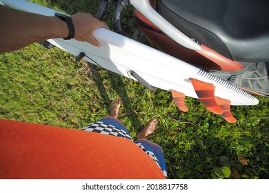 POV Shot Of A Man Surfer Wearing Blue Shorts And Orange T-shirt Take His White Short Surfboard From Scooter Moped.