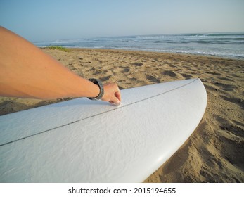 POV Shot Of Surfer Waxing Surfboard On Beach Next To The Ocean. 
