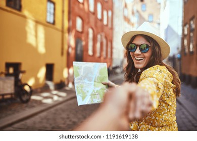 POV of a smiling young woman holding a map and wearing sunglasses leading another person by the hand while exploring city streets together