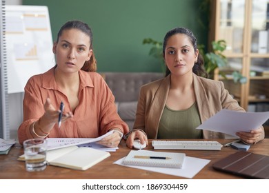 POV Of Two Businesswomen Looking At Camera And Pointing While Using Computer Sitting At Table In Office Against Green Wall