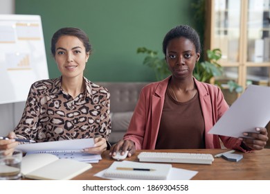 POV Of Two Businesswomen Looking At Camera And Using Computer While Sitting At Table In Office Against Green Wall