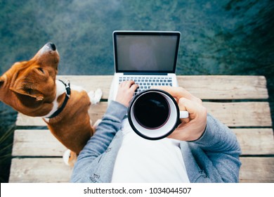 POV of urban millennial or hipster remote worker in startup work on laptop on sea, ocean or park lake wooden pier. Dog and best friend pet by side. Metal jug or cup with fresh coffee to stay awake