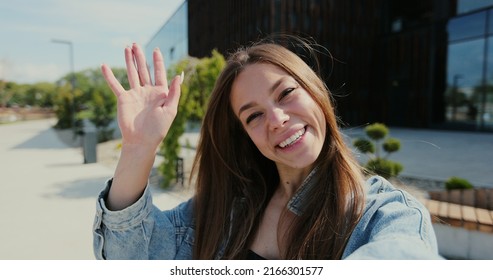 POV video call portrait of a young woman waving hand in casual clothes talking to friends while standing on city street.