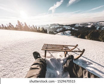 Pov View Of Young Man Looking The Sunset On Snow High Mountains With Vintage Sledding - Legs View Of Travel Influencer Enjoying Winter Landscape - Vacation Concept - Focus On Wood Luge