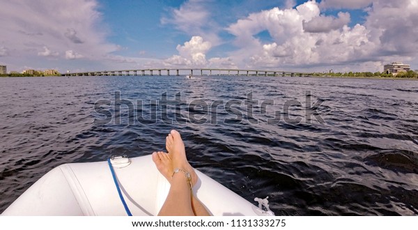 POV of a woman's crossed feet wearing a silver
anklet on a white inflatable dinghy boat traveling down a dark blue
river with a wide bridge and tall buildings under a bright blue,
cloud studded sky.