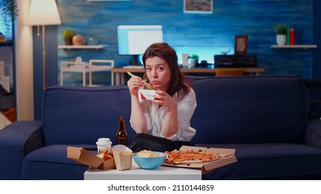 POV Of Young Woman Eating Chinese Food With Chopsticks While Watching Television After Work In Living Room. Adult Having Fast Food On Table Enjoying Asian Delivery Meal And Looking At Camera