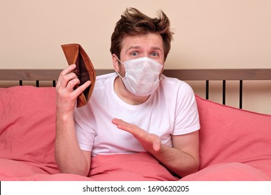 Poverty And Absence Of Money. Unemployed Sad Caucasian Man In Medical Mask Sitting At Home On Quarantine Or Isolation Because Of Covid Showing Empty Wallet. He Has No Money For Living.